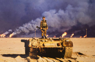 An American soldier stands on top of a destroyed Iraqi tank. Kuwaiti oil wells, ignited by Saddam's retreating forces, burn in the distance, Kuwait